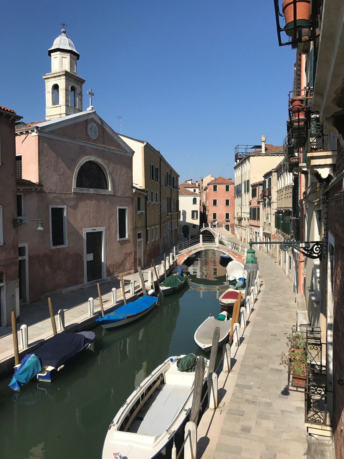 The image captures a serene canal scene, featuring colorful boats lining the waterway. Historic buildings with faded façades and a church steeple rise above the canal, reflecting local architectural styles. A small bridge connects both sides of the canal, enhancing the charm of the setting.