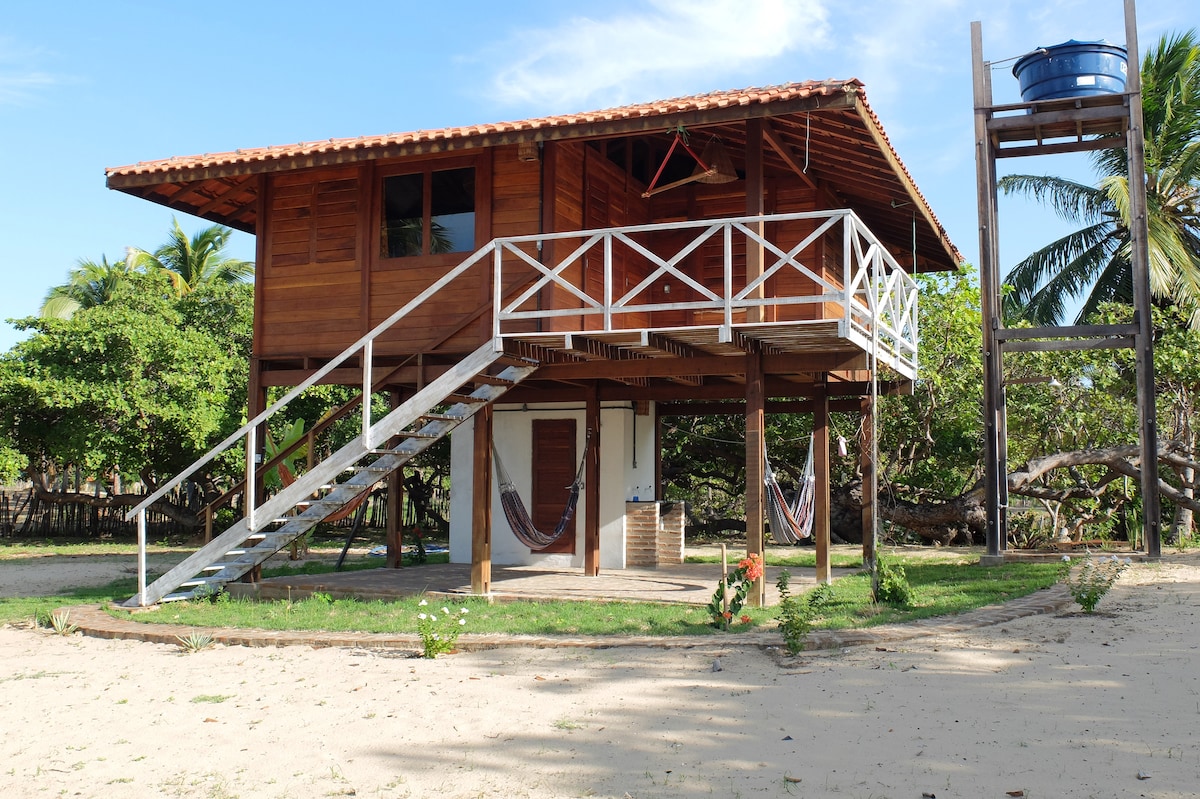 A wooden chalet stands elevated on stilt-like supports, featuring an exterior staircase leading to a wraparound deck. The building is surrounded by sandy areas and greenery, with hammocks visible underneath for relaxation.