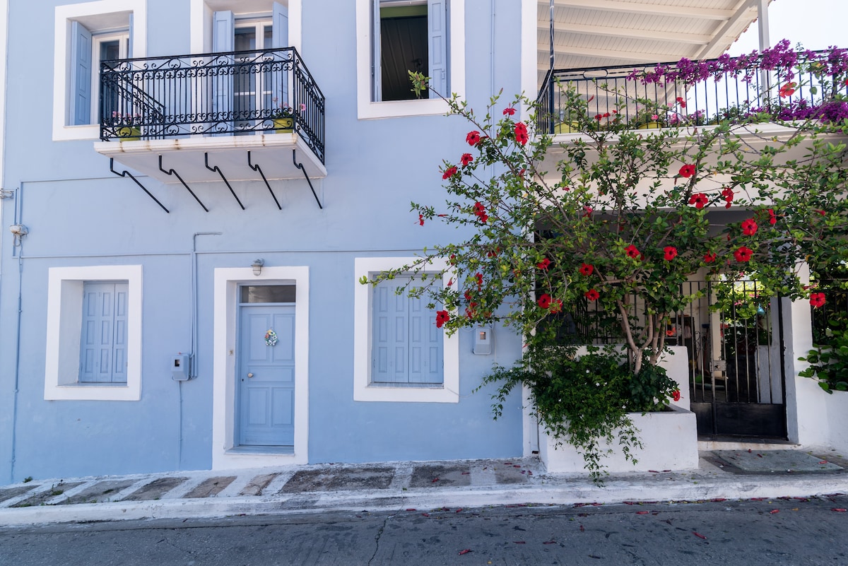 The exterior of the house is showcased, featuring a blue facade with white accents. Two balconies are adorned with potted flowers, while vibrant red blooms cascade from a nearby plant. A welcoming entrance is visible, with a light blue door and decorative wreath.