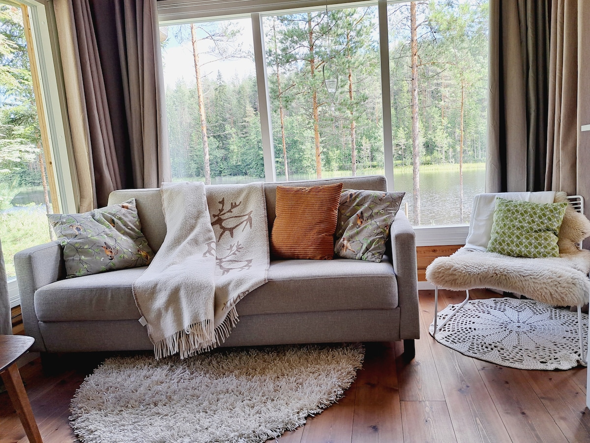 A comfortable living area is present, featuring a light gray sofa adorned with decorative pillows and a soft throw blanket. A white crochet chair complements the space, which is illuminated by natural light streaming through large windows overlooking a serene lake and forest.