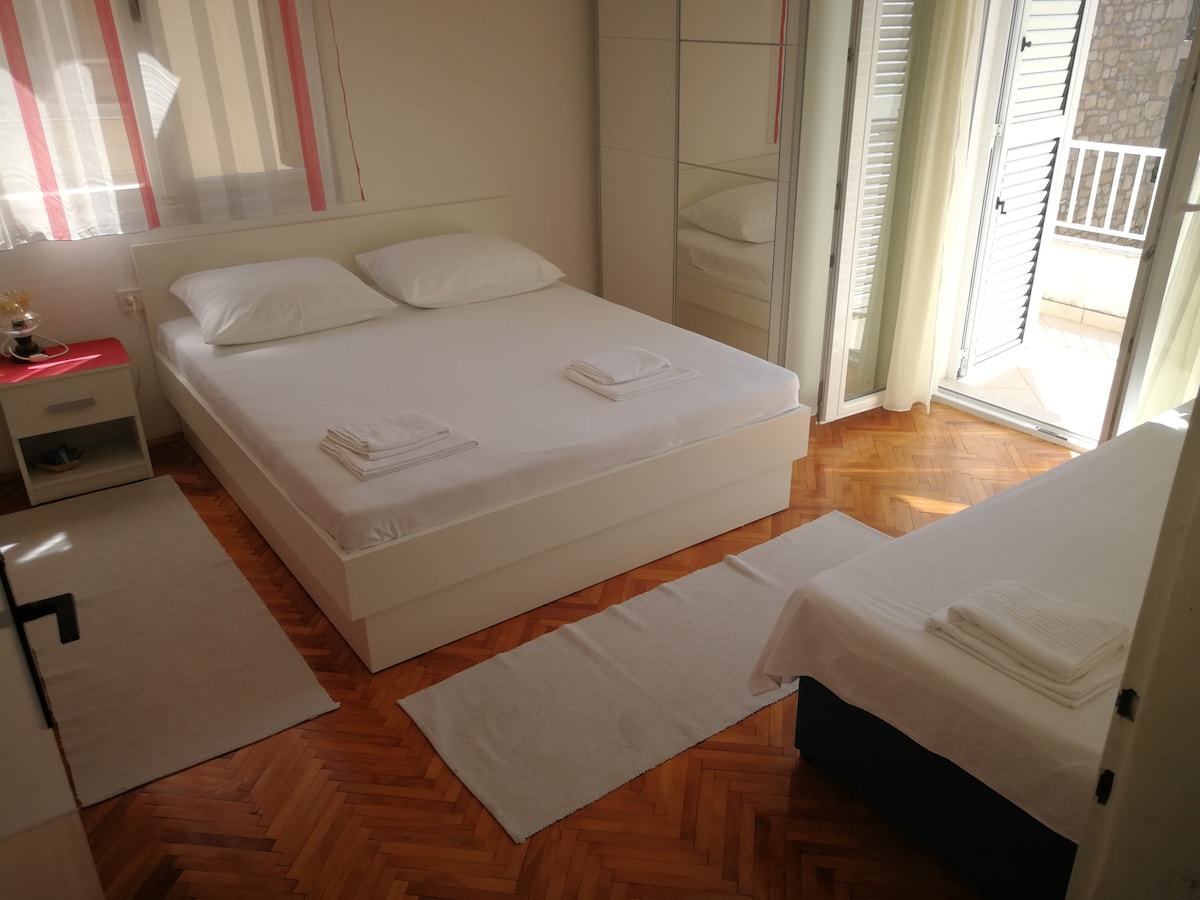 A bright sleeping area is presented with a spacious bed and a single bed, both dressed in crisp white linens. Natural light filters through the open glass doors, which lead out to a small balcony. The floor features light wood parquet and simple rugs.