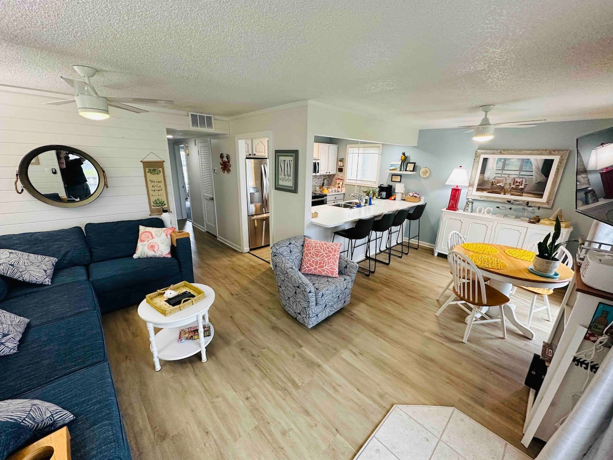 The open living area showcases a comfortable blue sectional sofa alongside a white side table. A dining space is present, featuring a round table with white chairs. The kitchen area is visible, highlighting modern appliances and a casual bar with additional seating.