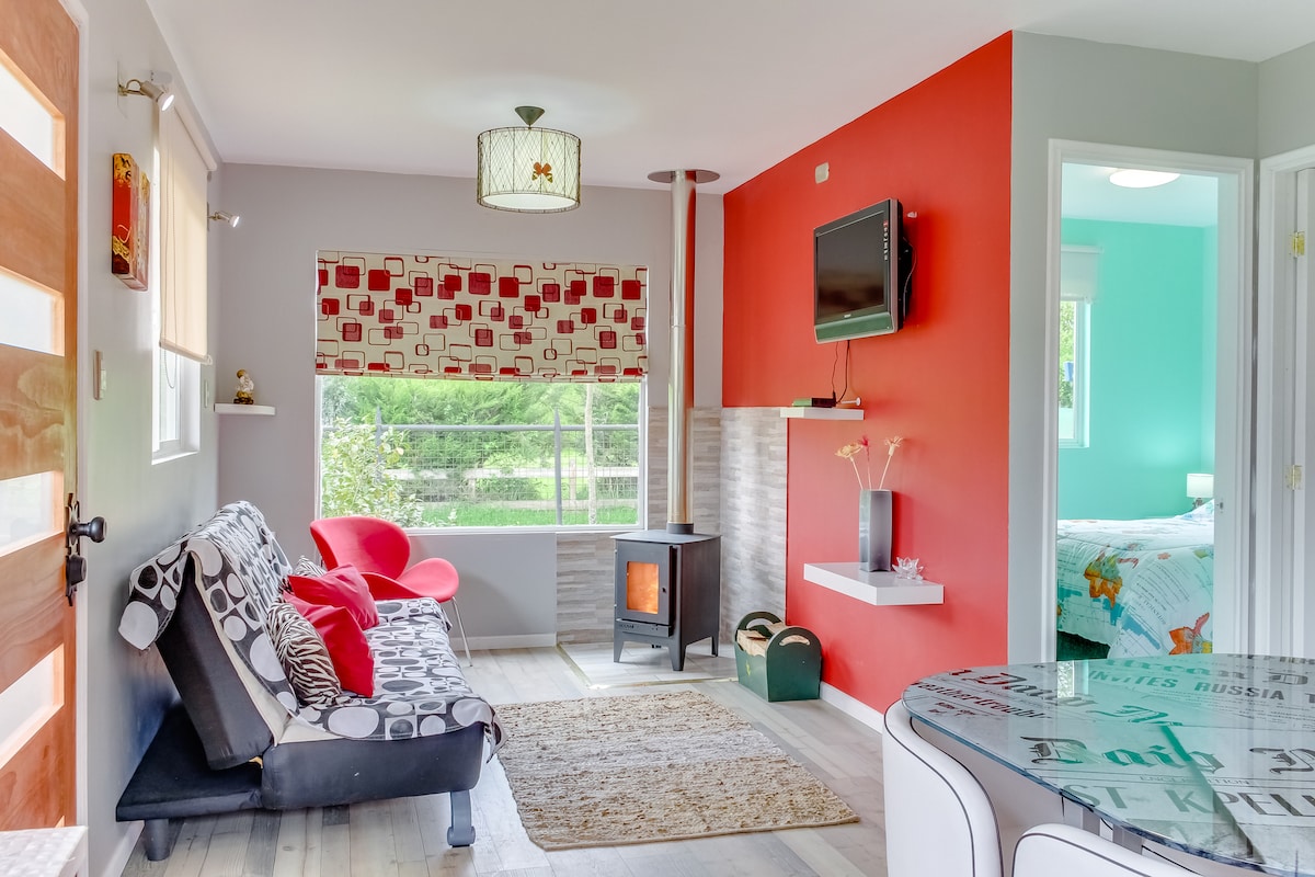 A bright living area features a comfortable sofa bed and a cozy wood stove, complemented by a flat-screen TV mounted on the wall. A round table is set nearby, and large windows provide natural light, showcasing views of the surrounding greenery.