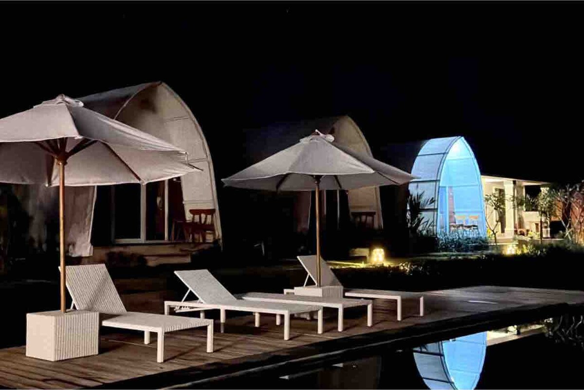 Lounge chairs are arranged beside a serene pool, accompanied by large umbrellas for shade. Structures with illuminated interiors are visible in the background, and the calm water reflects the night sky, creating a tranquil ambiance.
