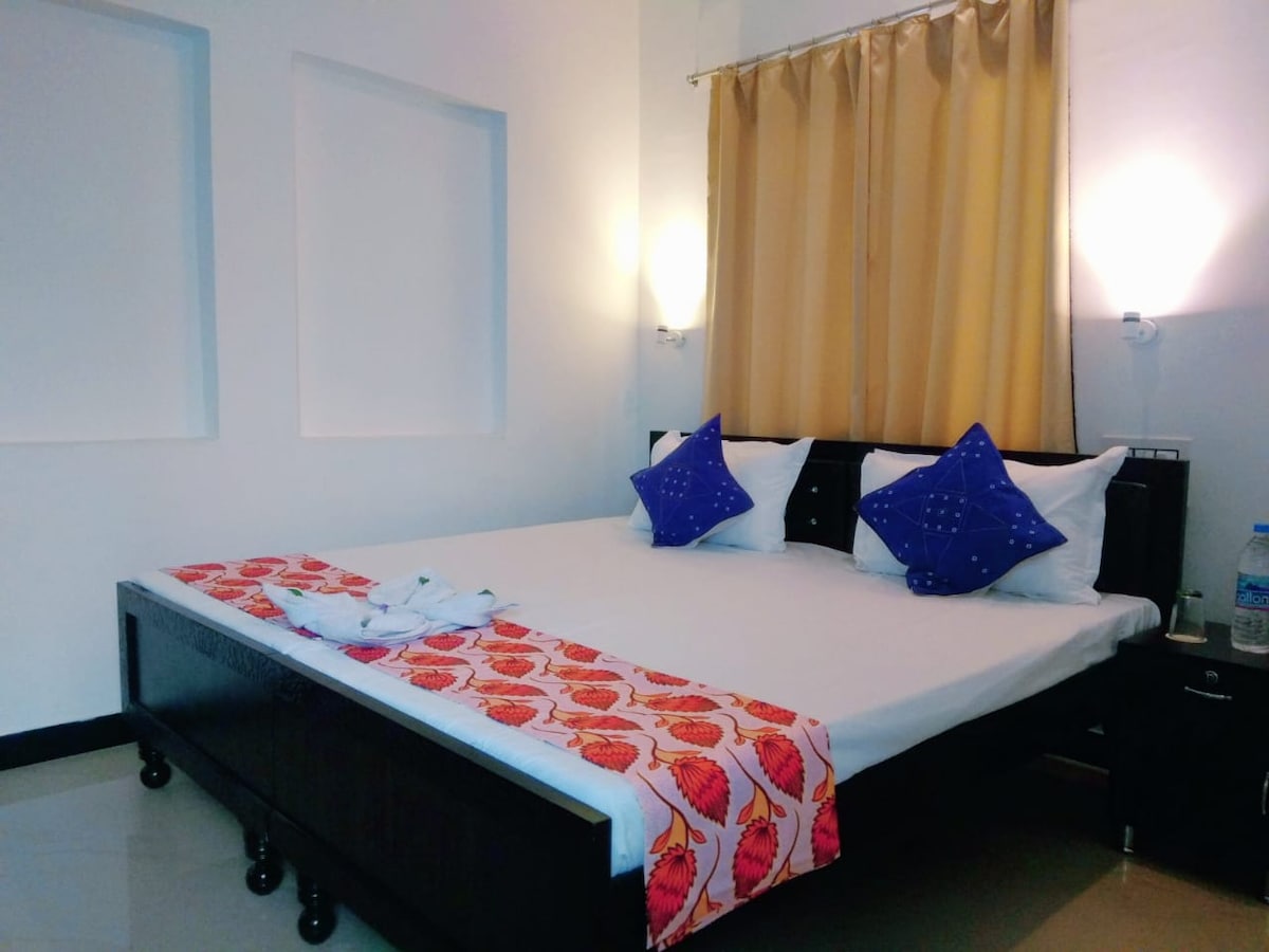 A spacious bed is set against a light-colored wall, adorned with blue accent pillows. The bedspread features a vibrant shell pattern, and warm light is provided by wall-mounted lamps on either side. A curtain in a soft hue filters natural light entering the room.