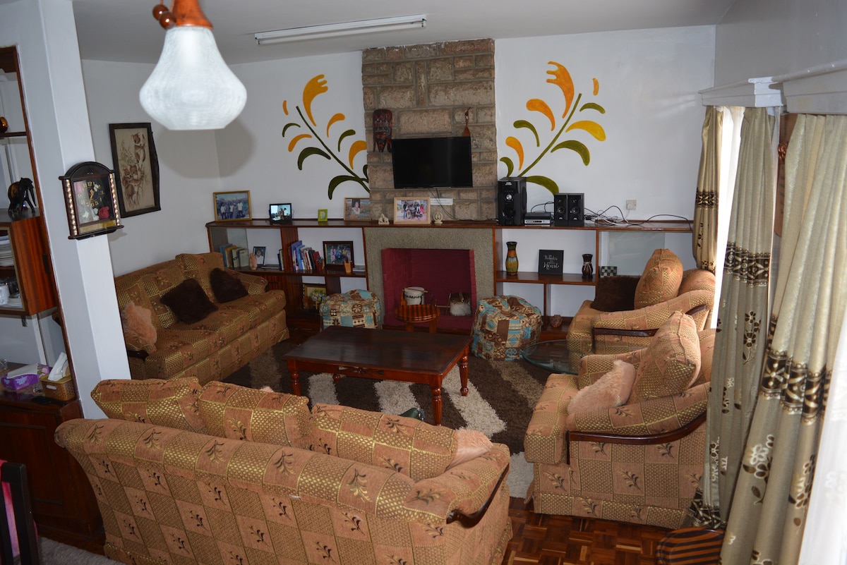 A spacious living area features a central wooden coffee table surrounded by a sectional sofa and armchairs. A stone fireplace is prominent, with a television mounted above. Shelving is visible in the background, displaying books and decorative items, while warm light filters through the window treatments.