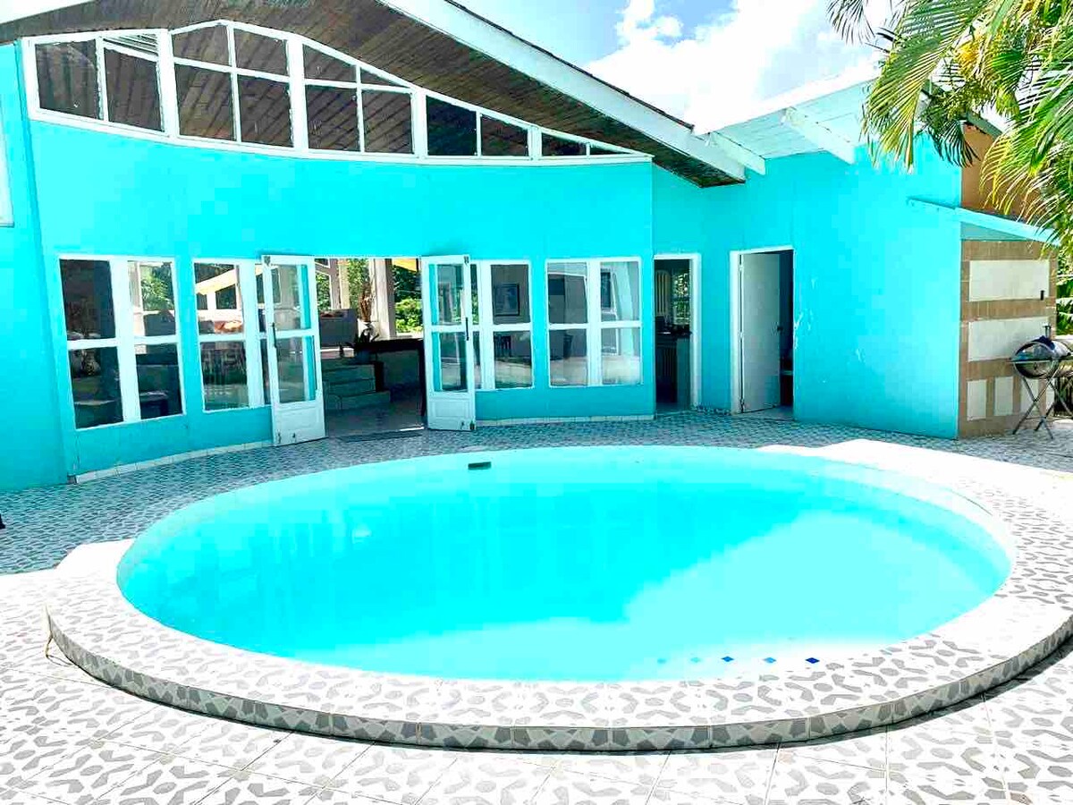 A private pool is surrounded by a tiled patio, reflecting a bright blue hue. Large glass doors lead into the villa, providing views of the inviting living space. Palm trees are visible in the background, adding to the tropical ambiance.