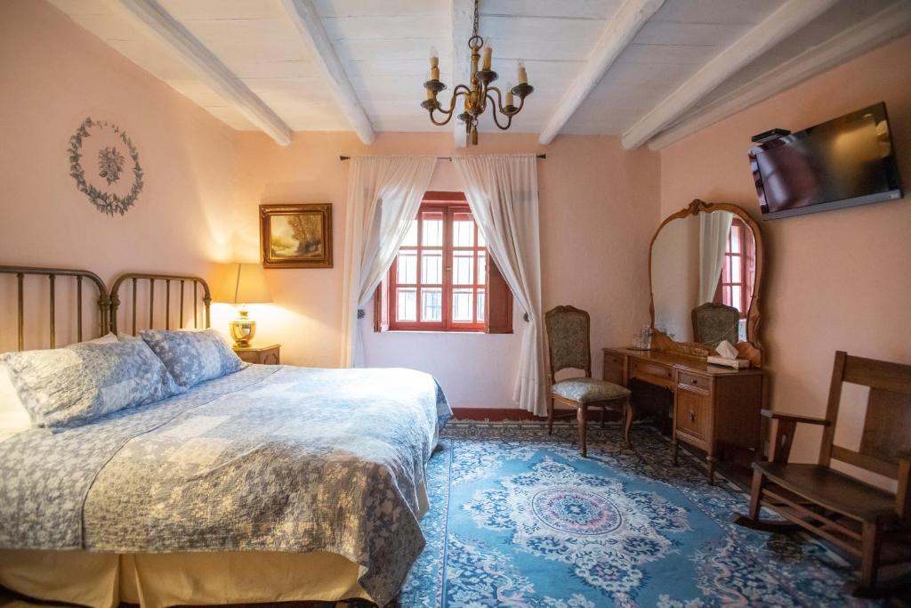 A cozy bedroom features a king-size bed with a patterned bedspread. A vintage chandelier hangs from the beamed ceiling, and light streams through two red-framed windows. A wooden rocking chair and a vanity with a mirror enhance the welcoming ambiance.