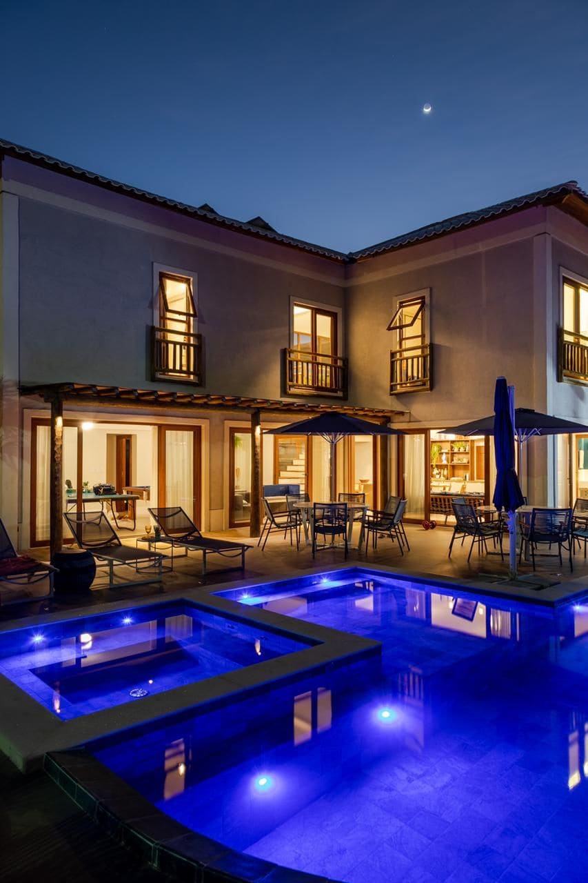 A spacious outdoor area is illuminated by soft lights reflecting off a large pool and a separate hot tub. Multiple lounge chairs surround the pool, and umbrellas provide shade. Large glass doors lead into the inviting interior of the house.