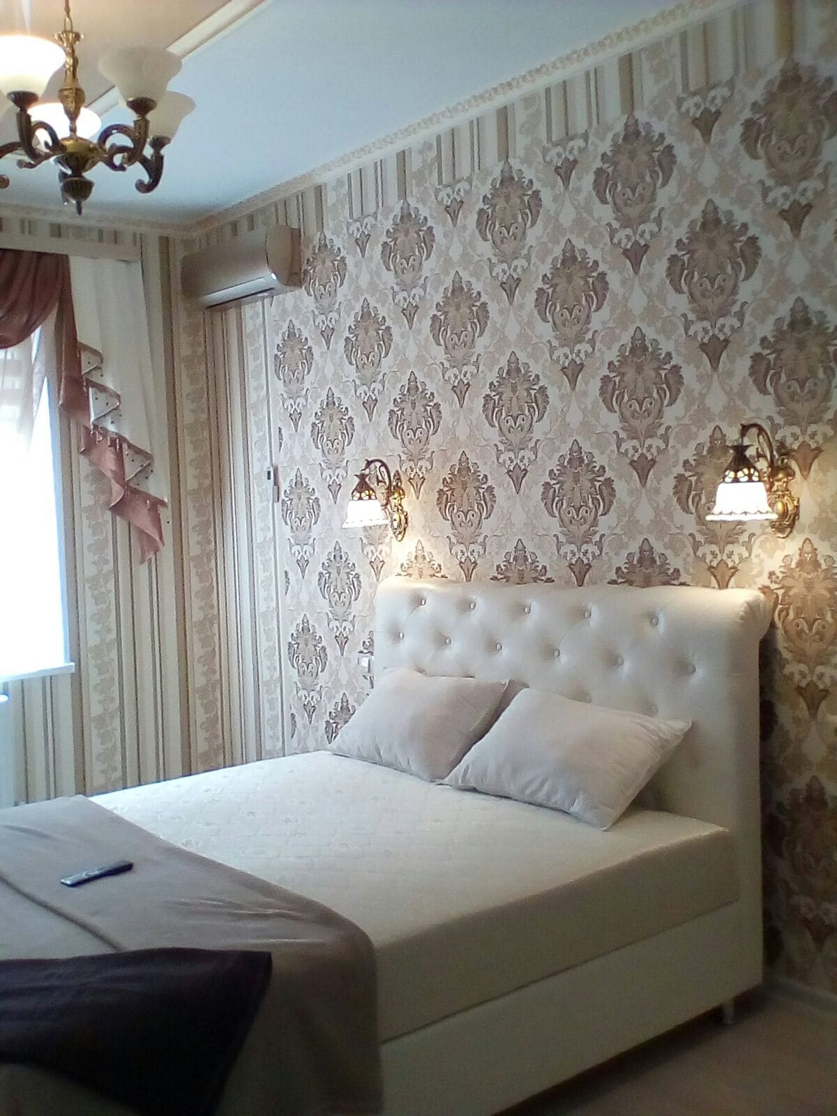 A cozy bedroom features a white, upholstered headboard with two decorative light fixtures on either side. Elegant wallpaper displays a floral pattern, while soft curtains frame a window. A neatly arranged bed with pillows invites rest and relaxation.