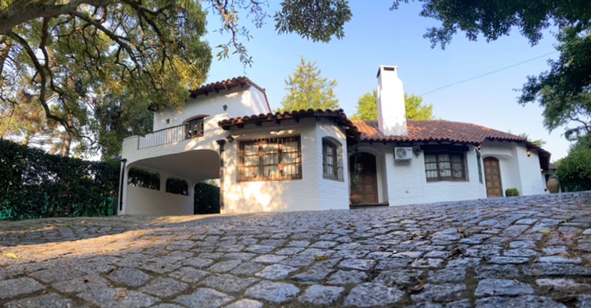 A charming colonial-style house is viewed from a cobblestone pathway, showcasing a white exterior with dark wooden accents. Lush trees provide shade, and a spacious balcony is visible above the entrance. The tranquil setting reflects the peaceful environment surrounding the home.