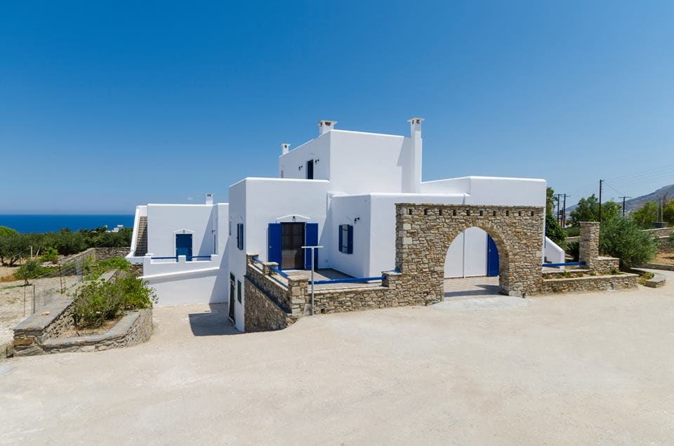 A modern villa is presented in a clean white façade with blue accents. A stone archway adds character to the entrance. The expansive grounds offer views of greenery and the sea in the background under a clear blue sky.