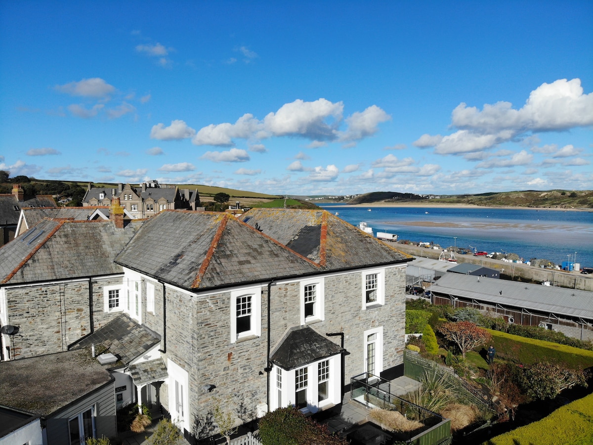Aerial view of No.1 Exbury, a renovated period property, showcases its stone exterior and spacious layout. The image captures the panoramic views of the serene Camel Estuary and nearby harbor, with a bright blue sky and scattered clouds enhancing the tranquil setting.