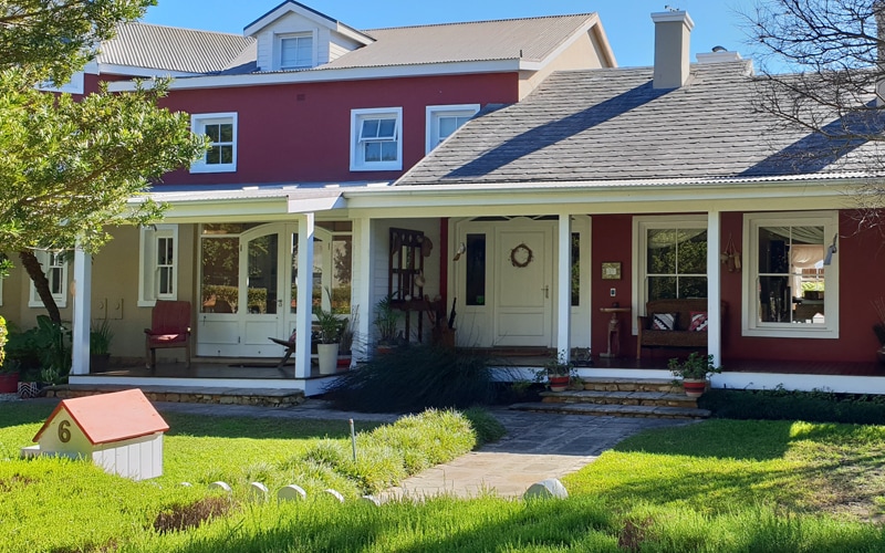 The exterior of the guest house displays a charming red façade complemented by white trim. A welcoming porch features multiple seating areas, and a decorative wreath hangs on the door. Lush greenery surrounds the pathways, enhancing the inviting atmosphere of the property.