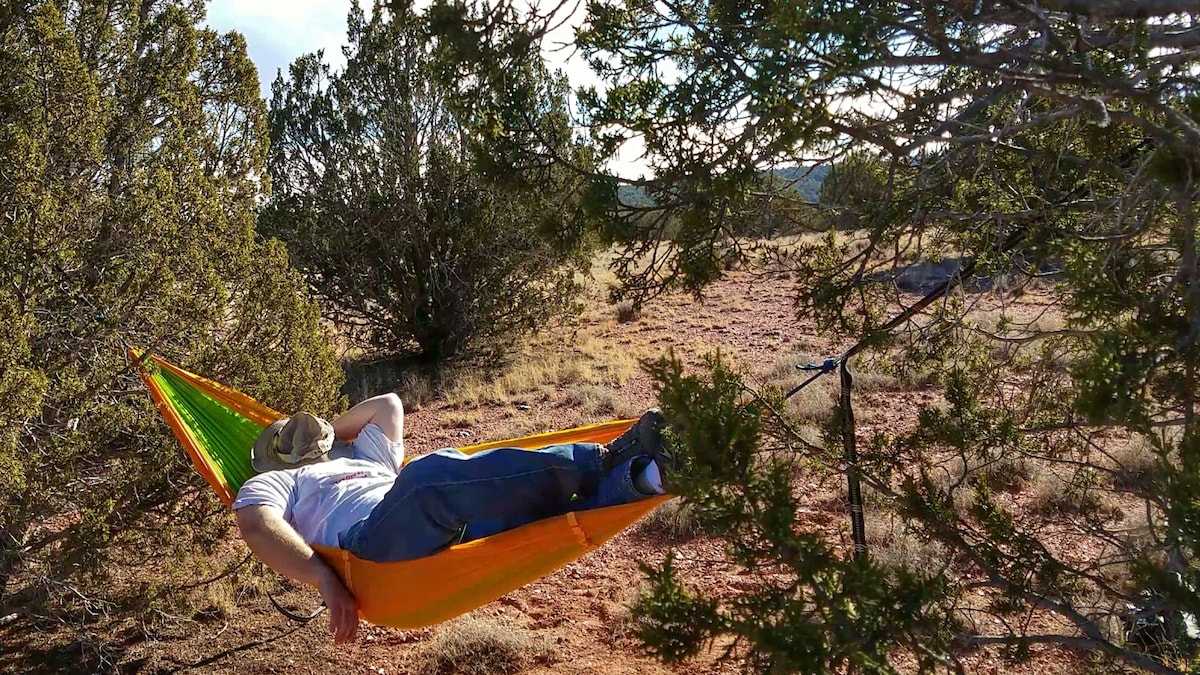 A colorful hammock is suspended between two trees in a serene outdoor setting. A person reclines comfortably, partially covered by a wide-brimmed hat. The surrounding landscape features gentle hills, sparse vegetation, and clear blue skies in the background.