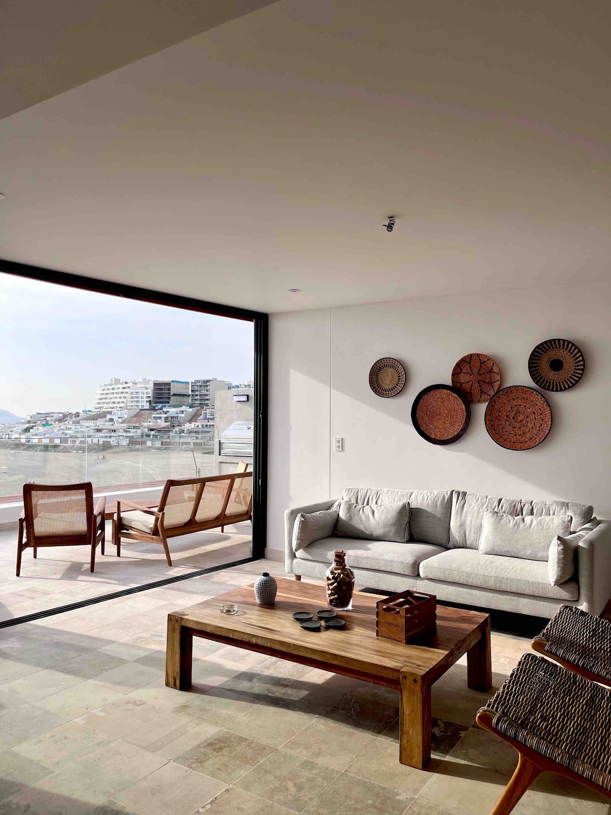 A light-filled living area features a comfortable gray sofa and a wooden coffee table with decorative elements. Wall art made of woven materials adds texture, while large glass doors offer views of the surroundings and access to an outdoor seating area.