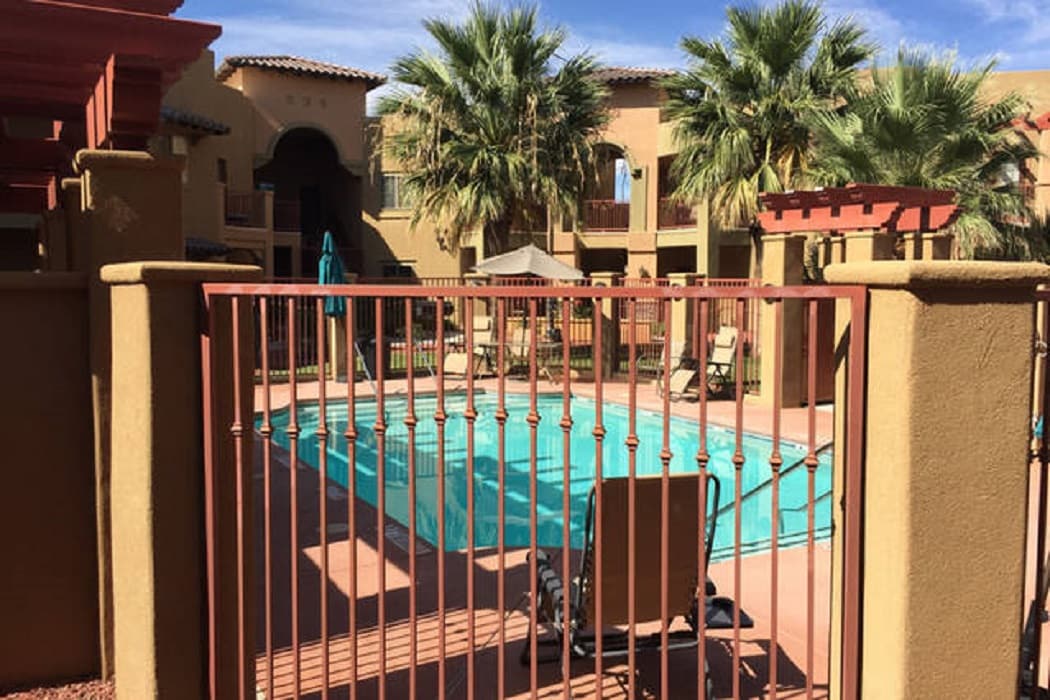 A fenced pool area features a clear blue swimming pool surrounded by paved decking. Palm trees and shaded seating areas provide a relaxing outdoor environment. Lounge chairs are situated at the edge of the pool, inviting for sunbathing or relaxation.