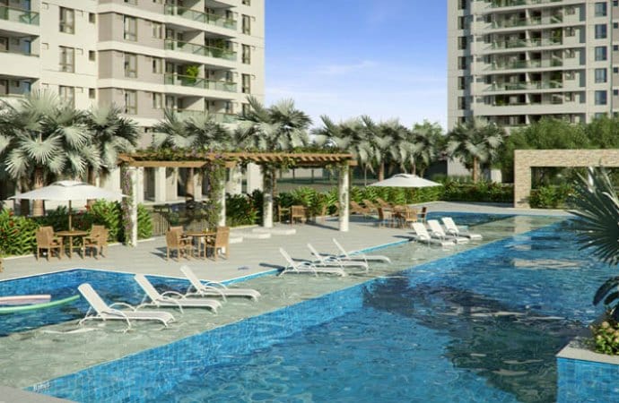 A swimming pool area is surrounded by landscaped gardens with palm trees and greenery. Lounge chairs are arranged along the poolside, and shaded seating areas with umbrellas are visible nearby. The modern buildings rise in the background under a blue sky.