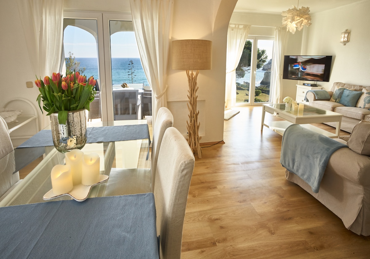A well-lit living area features a dining table adorned with a vase of fresh tulips and soft candles. Large glass doors provide a view of the sea, while a cozy seating arrangement with light-colored furnishings is visible. Natural light fills the space, enhancing the relaxed ambiance.