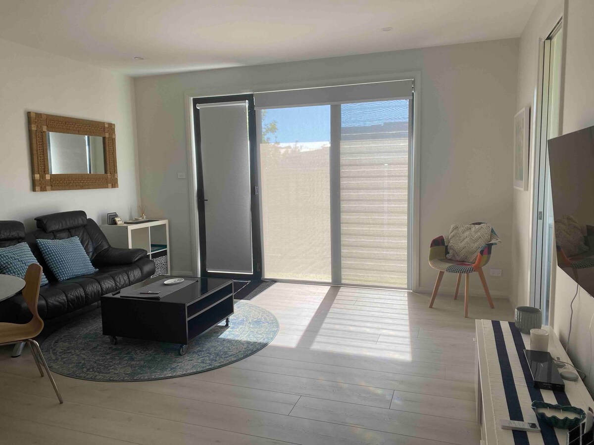 A spacious living area is highlighted by a black leather sofa adorned with decorative pillows. Natural light filters through large glass doors, complementing the light wood flooring. A round rug anchors the seating area, while a television is positioned on a low table opposite the couch.