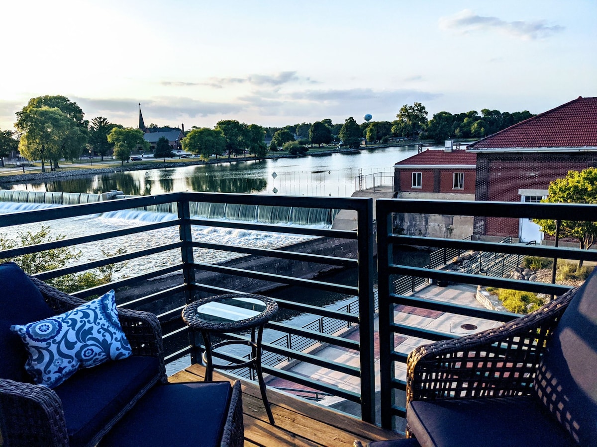 A cozy balcony features outdoor seating with cushions, providing a view of the Cedar River. The serene landscape includes trees along the shoreline and a backdrop of buildings, with gentle water flowing over a dam, illuminating the scene during dusk.