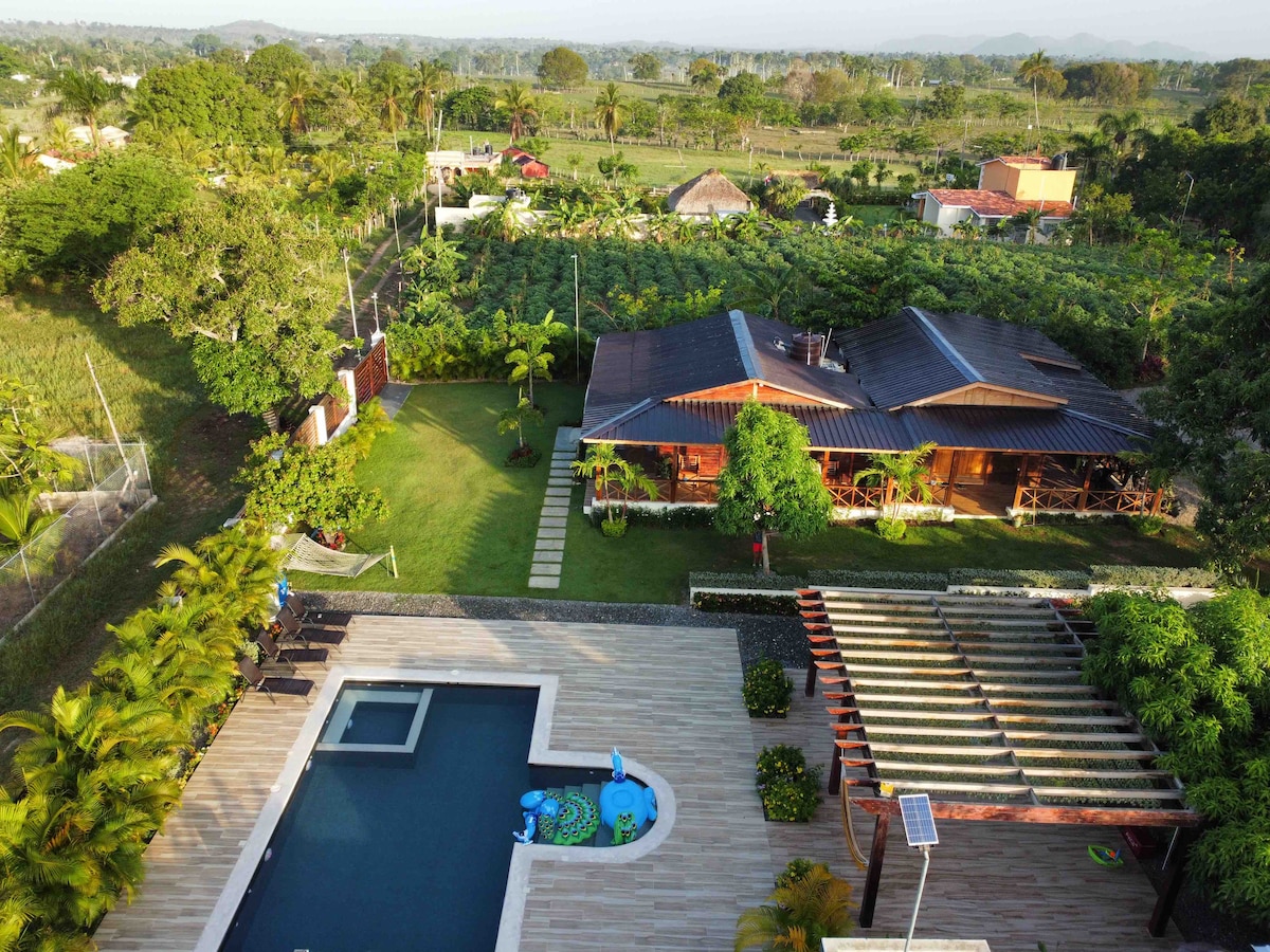 An aerial view showcases the property's spacious layout, featuring a clear swimming pool surrounded by lush greenery and well-maintained landscaping. Two wooden cottages are visible, with a natural path connecting them. The tranquil rural surroundings enhance the serene atmosphere of the cottage.