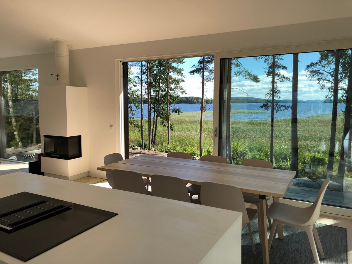 An open-concept dining area is presented, featuring a long wooden table surrounded by chairs. Floor-to-ceiling windows provide expansive views of the lake and surrounding greenery, allowing natural light to illuminate the space. A modern fireplace is visible within the living area.