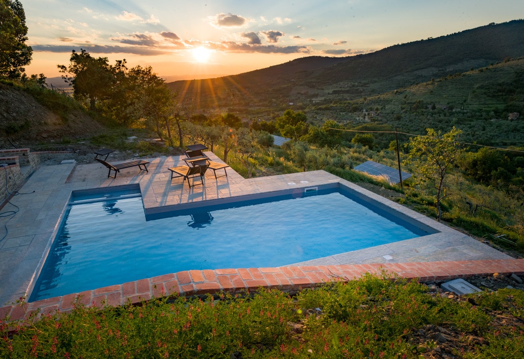A private swimming pool is situated amidst greenery, featuring a spacious deck with lounge chairs. The backdrop showcases rolling hills and a sunset that casts a warm glow over the landscape, creating a serene atmosphere.