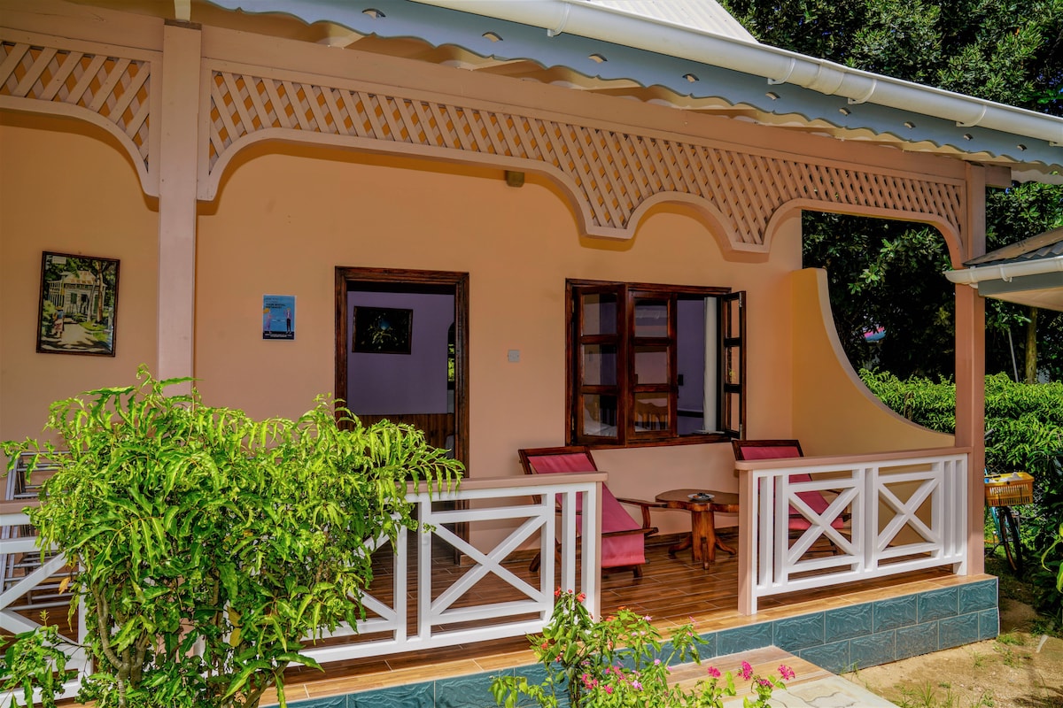 A charming porch with wooden furniture includes two red chairs and a small table, surrounded by lush greenery. The light-colored exterior displays decorative wooden elements, while windows framed in dark wood invite natural light into the space.