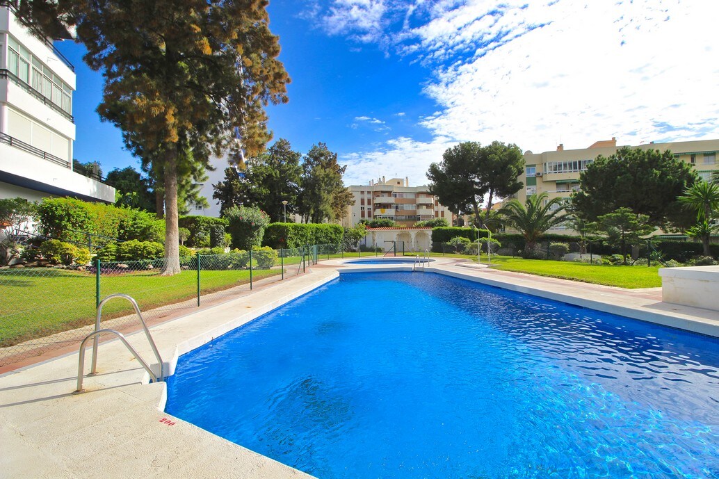 A spacious outdoor swimming pool is framed by lush greenery and well-maintained gardens. The water shimmers under the bright blue sky, with a ladder leading into the pool area. Sun loungers may be found nearby, providing a relaxing atmosphere for guests.
