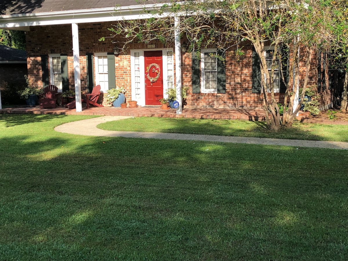 A welcoming facade of a brick home is framed by a well-maintained lawn. A curved pathway leads to a vibrant red front door, flanked by potted plants. The porch features wooden seating and is shaded by surrounding trees, creating a pleasant outdoor entry.