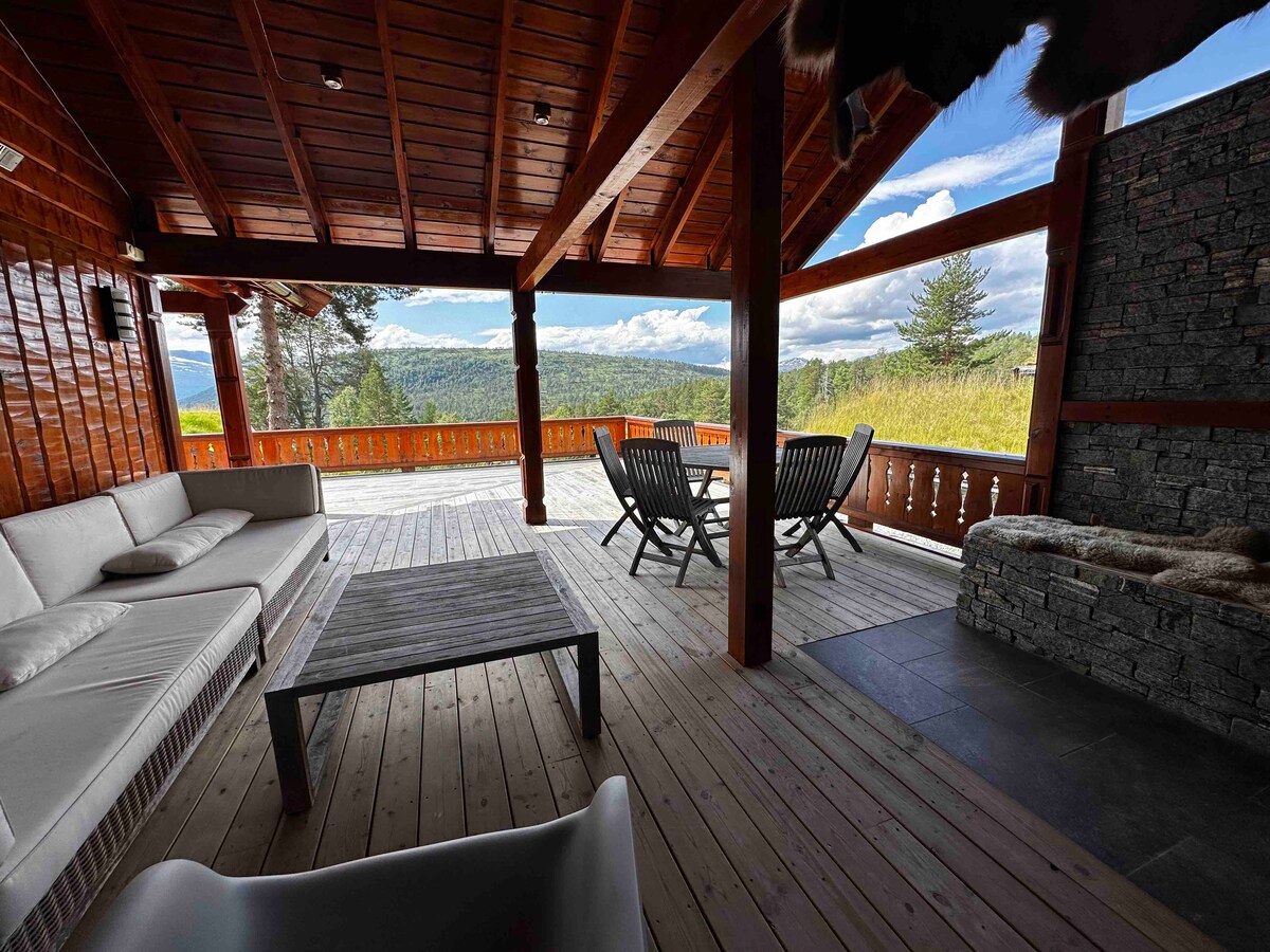 A spacious terrace features comfortable seating arrangements with a large sofa and lounge chairs, offering panoramic views of the surrounding landscape. A stone fireplace adds a cozy touch, while wooden beams enhance the rustic charm of the outdoor space.