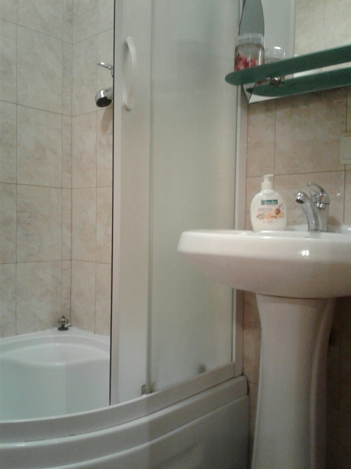 A compact bathroom features a glass-enclosed shower and a white pedestal sink. Light-colored tiled walls provide a clean backdrop, while a shelf holds personal care items, including soap. A bathtub can also be seen, positioned alongside the shower.