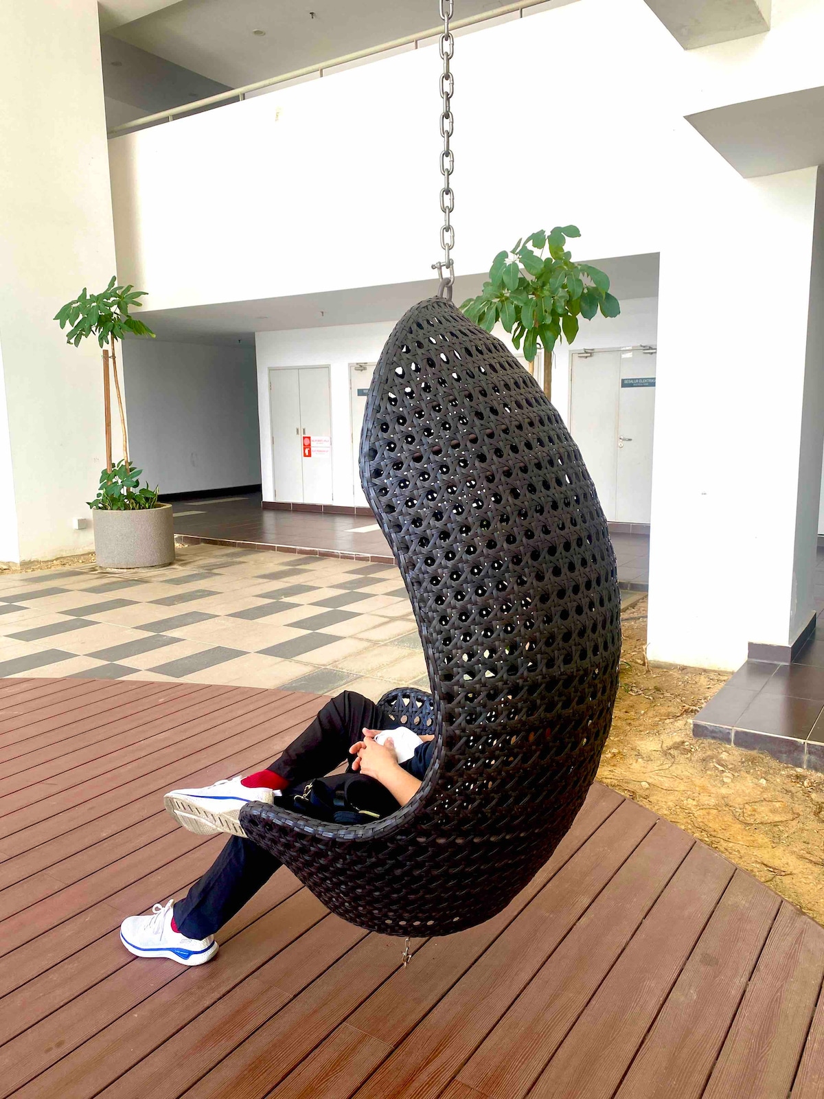 A unique hanging chair made of woven material is positioned on a wooden deck. Nearby, lush greenery is provided by potted plants, while a bright and airy space is created by the light streaming through large windows. The design encourages relaxation and comfort.