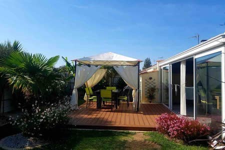 An outdoor terrace features a shaded seating area under a canopy, surrounded by greenery and colorful flowers. The space includes a table and chairs, providing a serene spot for relaxation. The adjoining glass structure allows natural light into the area.