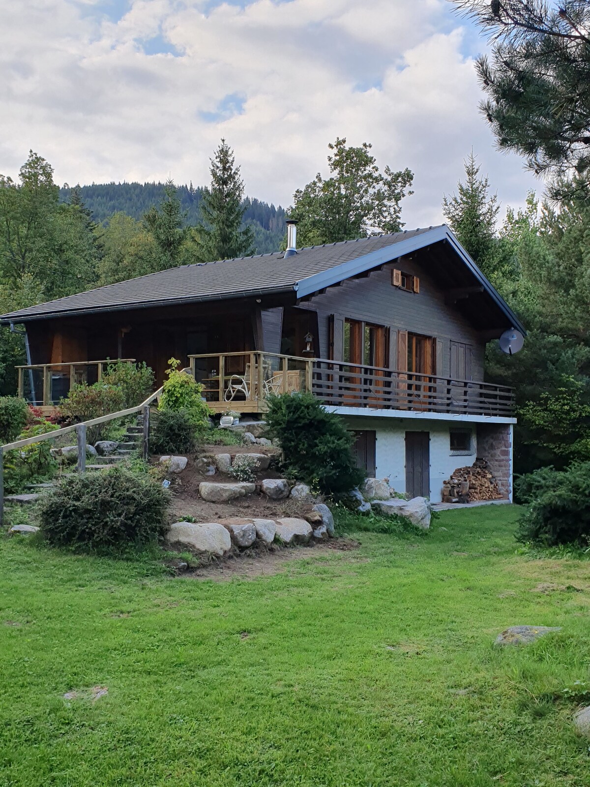 A chalet is nestled within a lush green landscape, featuring a wooden exterior and a spacious terrace. The structure is set against a backdrop of trees and mountains, with steps leading up to the entrance. A stacked wood supply is visible beside the house.