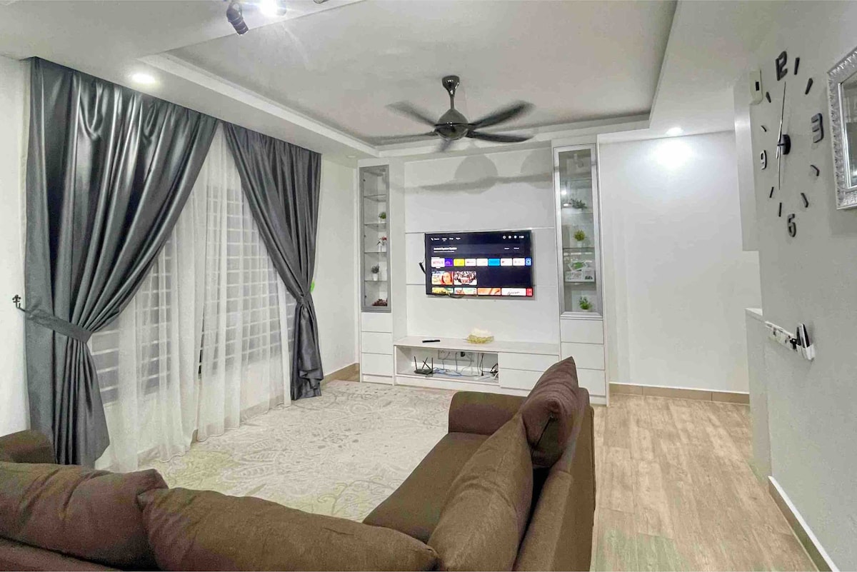 A spacious living area is equipped with a large wall-mounted television displaying various apps. Dark curtains frame the window, complemented by a cozy brown sofa. A ceiling fan provides airflow, and sleek shelving units enhance the minimalistic decor.