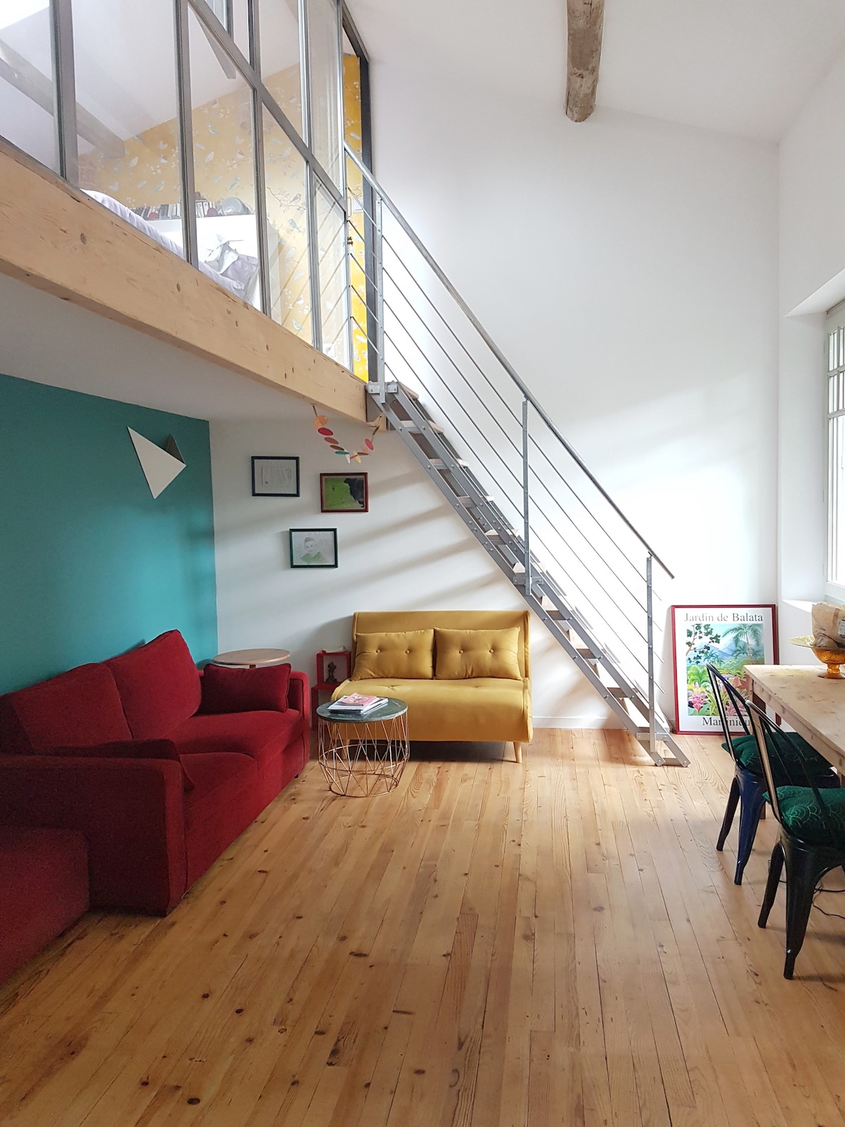 An airy living space is presented with high ceilings and wooden flooring. A red sofa and a yellow armchair are positioned near a small table. A staircase leads to a loft area, while framed artwork and colorful decor adorn the walls.