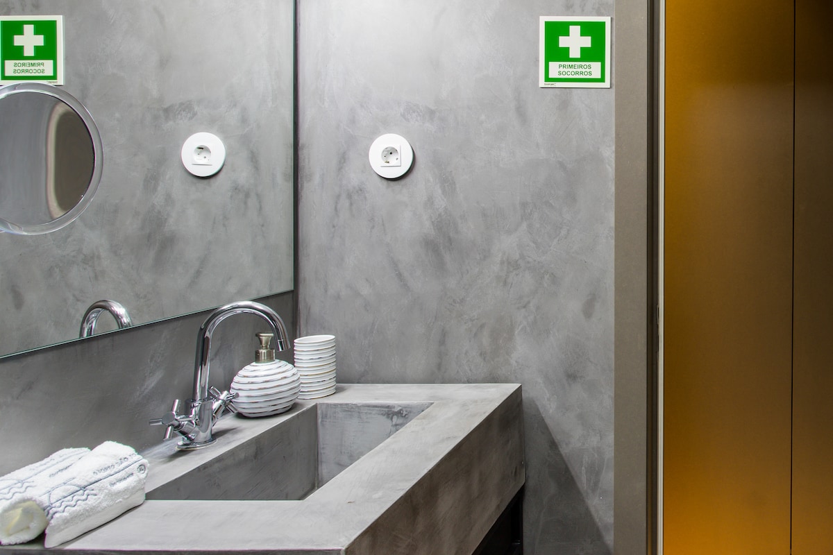The bathroom features a modern sink with a sleek stone countertop, complemented by a round mirror above. White towels and a set of plates are arranged neatly on the counter. Safety signage is displayed prominently, ensuring a secure environment.