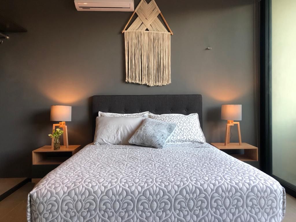 A spacious bed is positioned against a dark wall, surrounded by two nightstands with lamps providing gentle illumination. A textured wall hanging adds visual interest above the bed, while natural light enters through the large window, enhancing the serene ambiance.