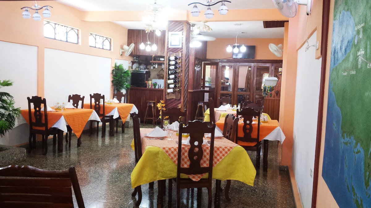 The dining area features several wooden tables, each adorned with checkered tablecloths in orange and yellow. Ceiling fans are visible, contributing to the relaxed atmosphere. A small bar area can be seen in the background, with a green wall depicting a map of the surrounding area.
