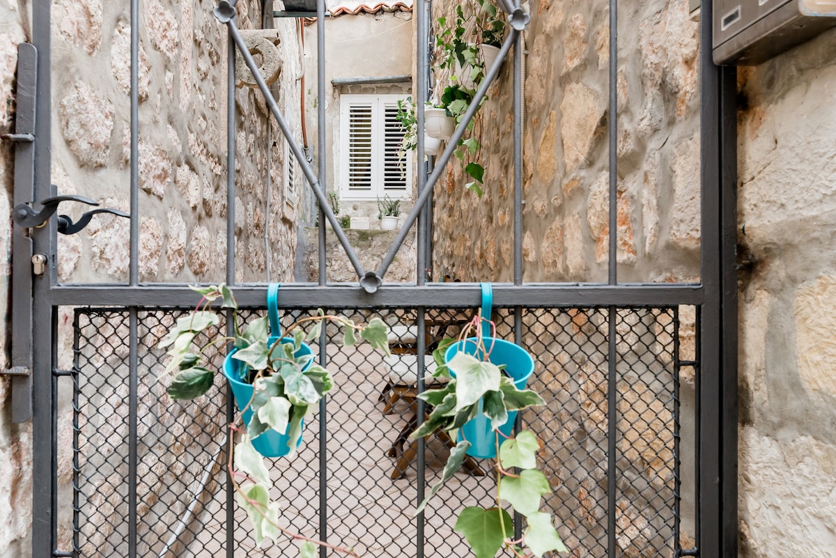 A gated entrance features a wrought iron design, creating an inviting entryway. Potted plants hang from the gate, adding a touch of greenery. The stone walls frame a narrow pathway leading to a secluded outdoor area with a wooden table, reflecting the charm of the surrounding environment.