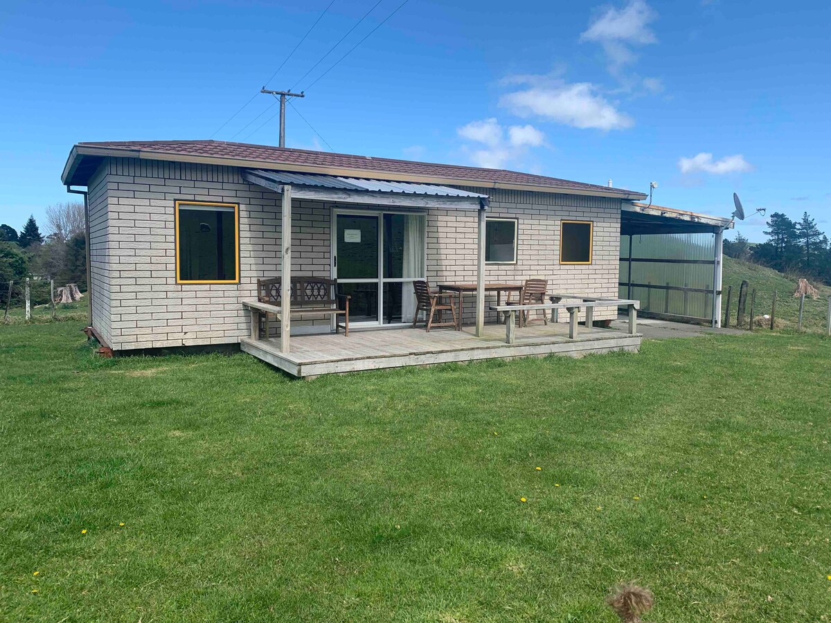 A single-story self-contained unit is situated on a grassy area, featuring a simple wooden deck with seating. Large windows allow natural light, while the exterior is composed of light-colored wooden panels. The property is surrounded by open land and greenery.