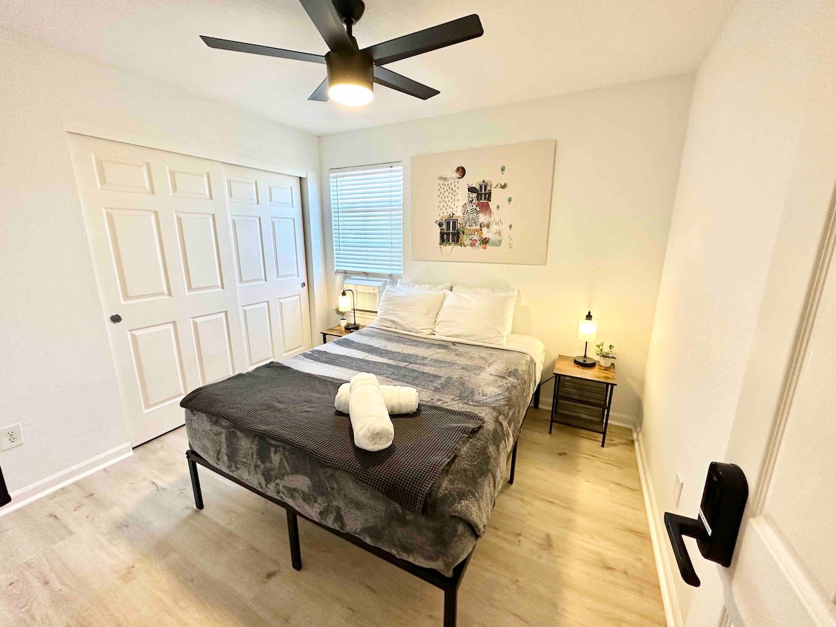 A bright and inviting bedroom features a spacious bed, complemented by soft linens and decorative pillows. A ceiling fan provides air circulation while natural light pours in through a window. Nightstands with lamps sit on either side of the bed, and a closet is visible in the background.