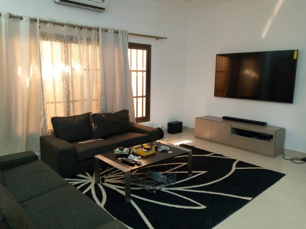 A comfortable living area is presented with a dark sectional sofa and a sleek coffee table. A large flat-screen TV is mounted on the wall, while natural light filters in through partially covered windows. A patterned area rug adds texture to the space.