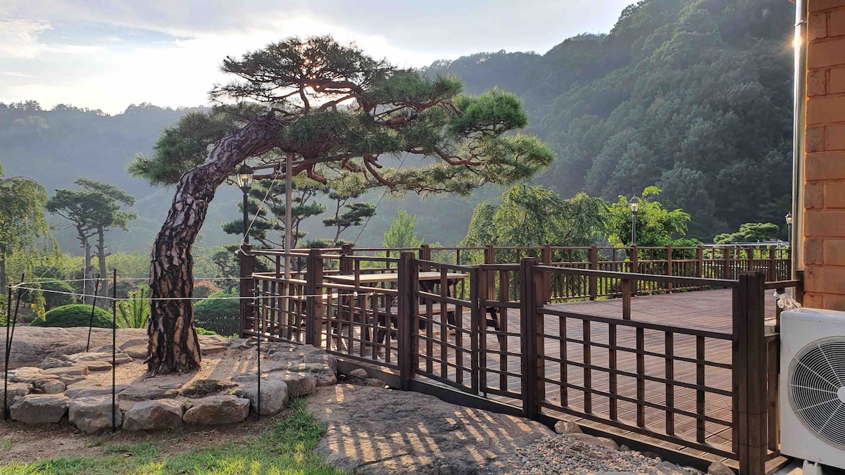 A spacious wooden deck extends into a natural landscape, framed by a twisted pine tree. Surrounding greenery contributes to a serene atmosphere, with distant hills and trees visible under soft daylight.