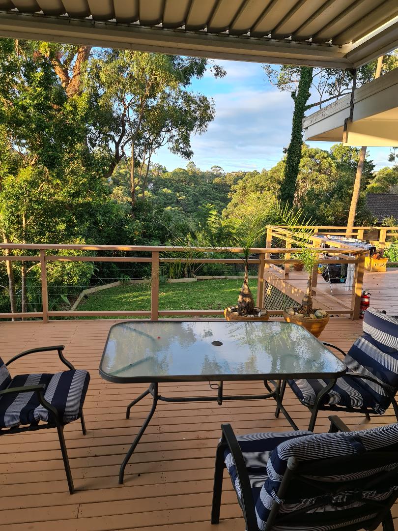 A deck area is seen with a glass-top table and two chairs, arranged to overlook a lush green landscape. Surrounding foliage creates a sense of tranquility, while natural light enhances the inviting atmosphere of this outdoor space.