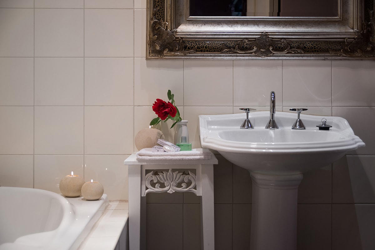 A bathroom featuring a classic pedestal sink with three chrome faucets. A decorative mirror with an ornate frame hangs above the sink. A small table holds a single red flower in a vase, accompanied by candles and toiletries, creating a serene and inviting ambiance.