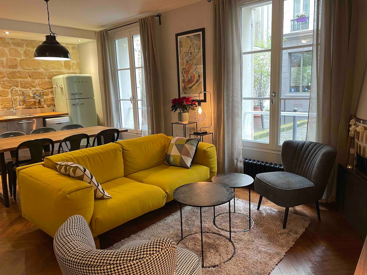 A bright living area features a yellow sectional sofa complemented by textured chairs in various patterns. Two small tables are positioned on a soft area rug. Natural light enters through large windows, revealing a glimpse of the courtyard outside.