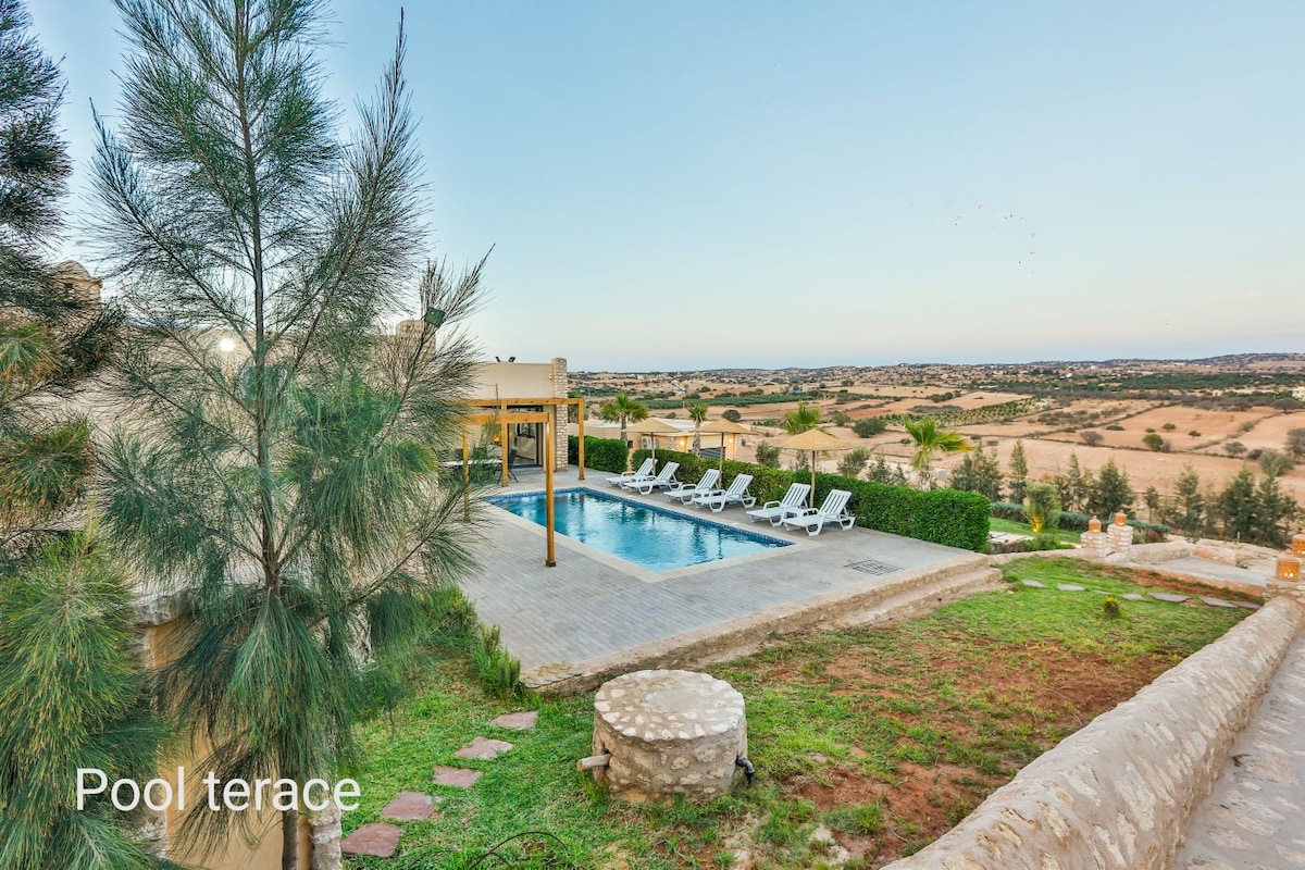A clear view of the pool terrace showcases a private swimming pool surrounded by loungers, set against a backdrop of rolling hills. Lush greenery frames the area, emphasizing tranquil outdoor living in a serene landscape.